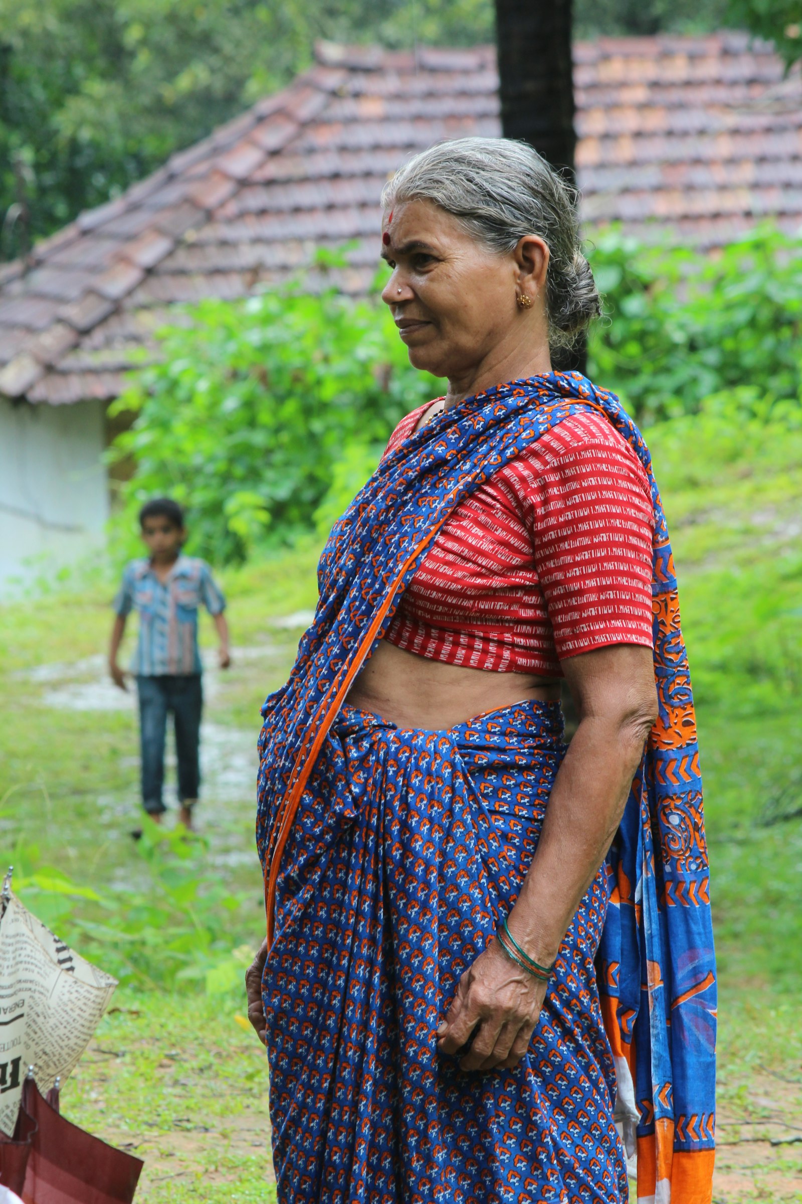 An older woman in a sari outside her home in a village, a child behind her