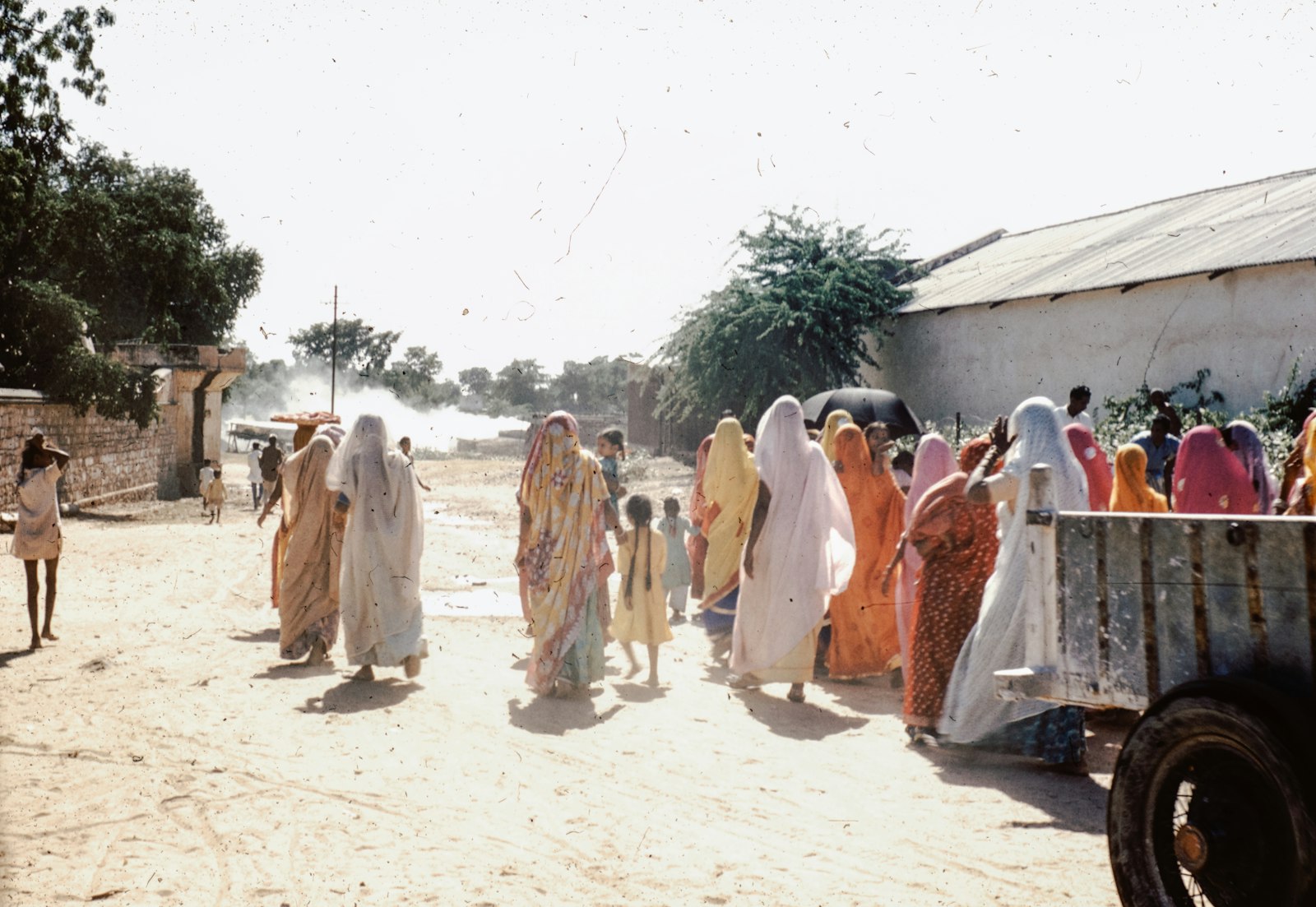 Women in saris walking down a sunlit village road toward a gathering