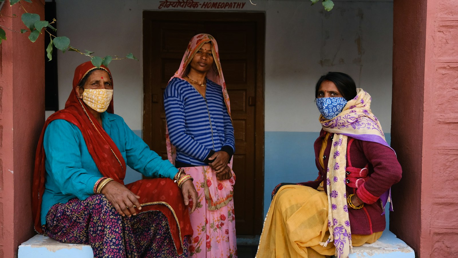 Three women seated outside a village home, two wearing masks